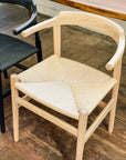 A SOBU Thor Dining Chair - White Ash, with a solid ash frame and natural woven paper cord seat, sits on a wooden floor next to a black chair of similar design.