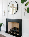 A modern fireplace with a matte black finish and herringbone brick interior sits against a white paneled wall. Above the mantel, the SOBU Toledo Mirror - Round hangs between wall sconces, with green plants nearby.