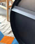 Close-up of the SOBU Toledo Mirror - Round with a matte black finish, shown on its side. Part of a wooden chair and a geometric rug in orange, blue, and beige are visible in the background.
