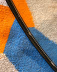 Close-up of the SOBU Toledo Mirror - Round, featuring a matte black rim, displayed diagonally on a grey carpet with bold orange, blue, and light blue geometric patterns.
