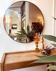 A wooden table with a decorative plant, dried flowers, and a wooden figurine sits beneath the SOBU Toledo Mirror - Round, featuring a matte black steel frame and reflecting greenery and part of the rooms interior.