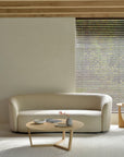 A minimalist living room with a curved beige sofa and the Ethnicraft Tripod Coffee Table - Oak, topped with two ceramic vessels and a magazine, complemented by wooden blinds letting in natural light and classic European refinement.