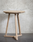 The Ethnicraft Tripod Side Table - Oak features a small, round European oak top with three angled legs, set against a textured concrete wall and dark floor—highlighting the brands signature elegant design.