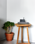 The Ethnicraft Tripod Side Table - Teak displays folded gray towels and a black device next to a potted green plant, set against a white wall in a minimalist room.