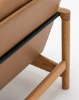 Close-up of the Troubadour Lounge Chair by Wooden Stories, featuring light oak legs and frame, a tan leather seat, and a sleek black metal support bar, set against a plain white background.