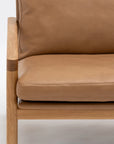 Close-up of the Wooden Stories Troubadour Lounge Chair | Leather • Light Oak, showcasing its solid light oak frame and tan leather cushions with focus on the armrest, seat, and backrest against a white background.