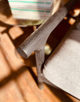 Close-up of the SOBU Viking Lounge Chair - Black Ash’s wooden armrest on gray upholstery, featuring a solid ash wood frame. A round glass table with magazines sits in the background as sunlight casts shadows on the wooden floor.