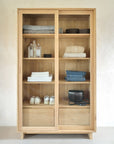 The Wave Storage Cupboard - Oak by Ethnicraft, crafted from European oak with sliding glass doors, is arranged against a white wall and displays towels, clothes, baskets, toiletry bottles, shoes, and a black bag on its shelves.