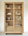 The Ethnicraft Wave Storage Cupboard in oak features sliding glass doors and is neatly arranged with books and minimalistic white decor like vases, bowls, and a sculpture on its shelves.