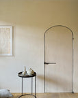 Minimalist room with a light-colored arched door, an Ethnicraft round black side table topped with the White Slice Wooden Tray (Round/Small), a teapot and vases, abstract art on the wall, and two gray floor cushions on wooden flooring.