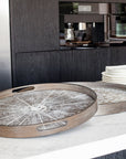 Two small, round White Slice wooden trays by Ethnicraft rest on a white marble countertop, with stacked square plates and salt and pepper shakers nearby and a modern black kitchen in the background.