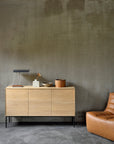 Minimalist living room with the Ethnicraft Whitebird Sideboard - 3 door adorned by a lamp, dried flower vase, bowl, and wooden container. A tan leather chair sits beside a textured gray wall and concrete floor.