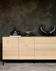 The Ethnicraft Whitebird Sideboard - 71 in solid oak with black metal legs stands against a dark textured wall, topped with a black vase, rustic bowl, and black horn-shaped sculpture.