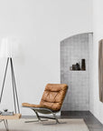 A modern minimalist room featuring a brown leather lounge chair, tripod floor lamp with white shade, low wooden coffee table, and the Armadillo&Co Winnow Rug in Alabaster adding warmth, with decorative vases in a niche against a tiled wall.