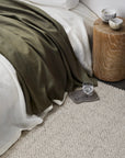 A cozy bed with white sheets and an olive green blanket sits beside a wooden stump nightstand with a bowl and cup. A glass of water rests on a tray atop the Winnow Rug - Alabaster by Armadillo&Co.