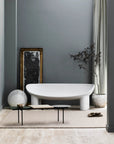 Minimalist bathroom with a modern white bathtub, black and gold mirror, potted plant, round textured lamp, low black table with decor, slippers, and a neutral Armadillo&Co Winnow Rug - Granite made from 100% wool.