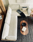 A top-down view of a modern living room showcasing SOBU’s Woody Sectional Sofa in White Ash and Fog, paired with a wooden oval coffee table displaying a black bowl and books atop a dark textured rug.