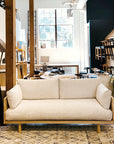 The SOBU Woody Sofa SAMPLE_(DZ), featuring a light hue and solid ash wood frame, is displayed in a bright, contemporary showroom with large windows, stylish furnishings, and a patterned rug placed in front of the sofa.