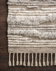 The Loloi Khalid Rug in Natural/Ivory features Moroccan-inspired patterns and raised, shaggy textures with fringe tassels on one edge, adding visual interest to any dark wooden floor.