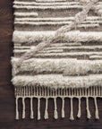 A close-up of the Loloi Khalid Rug in Ivory/Taupe shows cream and brown stripes, textured patterns, and braided tassels along the edge. The hand-knotted 100% wool rug is displayed on a dark wooden floor.