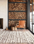 A modern living room features a large wall-mounted firewood rack, a tan leather chair, a small wooden stump table, and the Loloi Khalid Rug in Ivory/Taupe. Glass doors open to a wooden deck, enhancing the Moroccan-inspired ambiance.