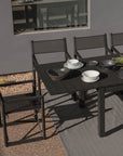A modern outdoor dining table is paired with four Harbour Pacific Folding Chairs in black teak, set with white and black ceramic plates and bowls. The table rests on a textured rug beside a gray wall with a window and gravel surroundings.