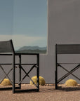 Two Harbour Pacific Folding Chairs in black teak sit on gravel by a gray building with a large window, surrounded by round cacti. The window reflects mountains, creating a stylish outdoor setting.