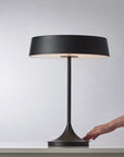The Seed Lighting China LED Table Lamp - Black, featuring a modern matte steel base and wide circular shade, rests on a white surface. A hand reaches for its dimmable spin base to control the LED. The background is light gray.