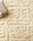 The Loloi Hygge Rug in Oatmeal/Sand, hand-loomed from 100% wool with a textured geometric design, is styled with a neutral book and a white knot ornament on one corner.