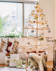 The SOBU Immortal Christmas Tree, a minimalist and sustainable wooden design, is decorated with ornaments and string lights, standing by the window among wrapped gifts, stockings, and cozy fur throws in a bright festive room.