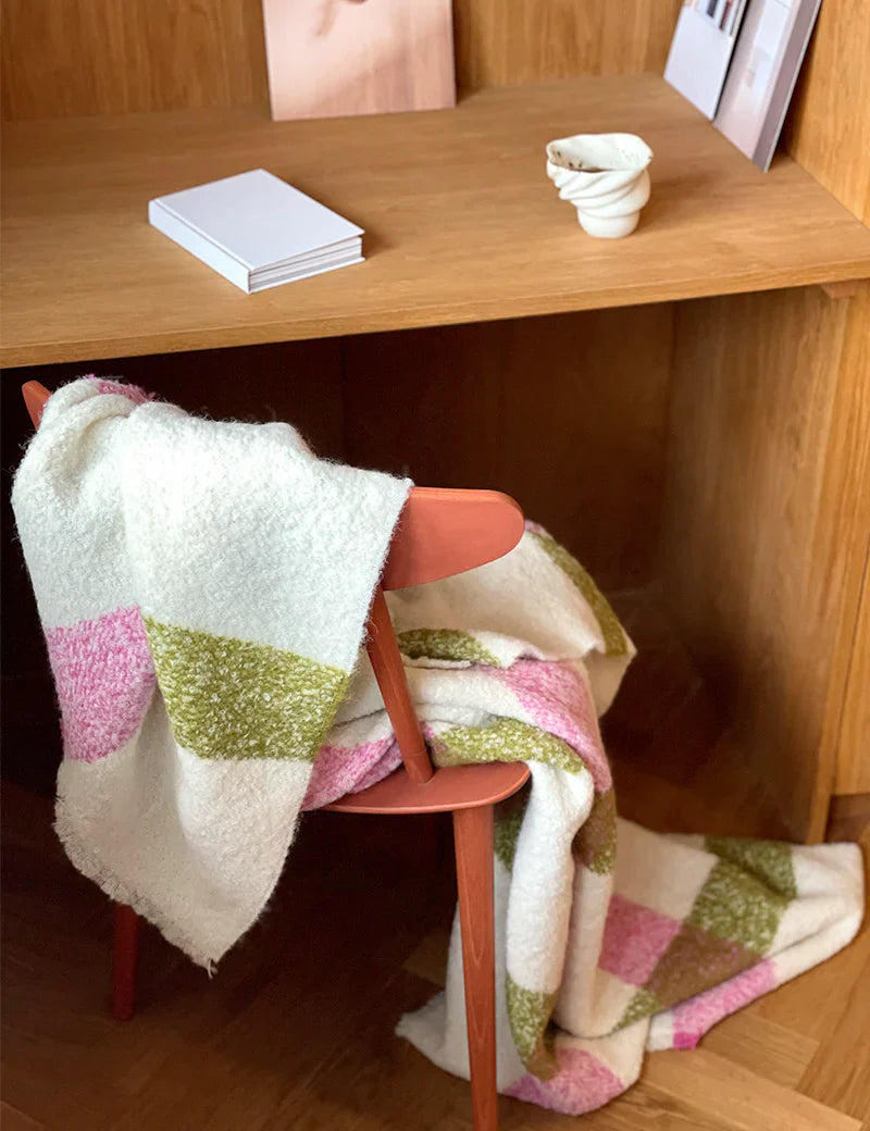 A soft, white blanket with green and pink stripes draped over a wooden chair, set in front of a wooden desk with notebooks and a small ceramic cup on top.