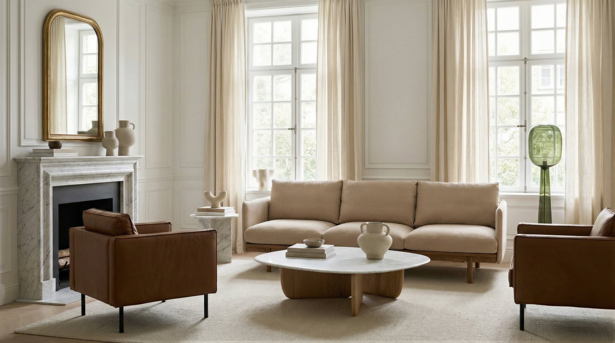 A modern living room with a beige sofa, two brown armchairs, a marble coffee table, a fireplace with a mirror above, large windows, cream curtains, and neutral-toned decor.