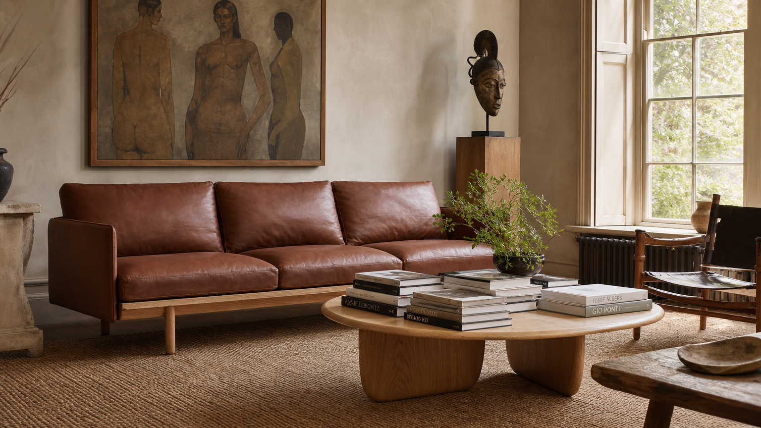 A modern living room with a brown leather sofa, wooden coffee table with books, a sculpture, a large painting, and a window letting in natural light.