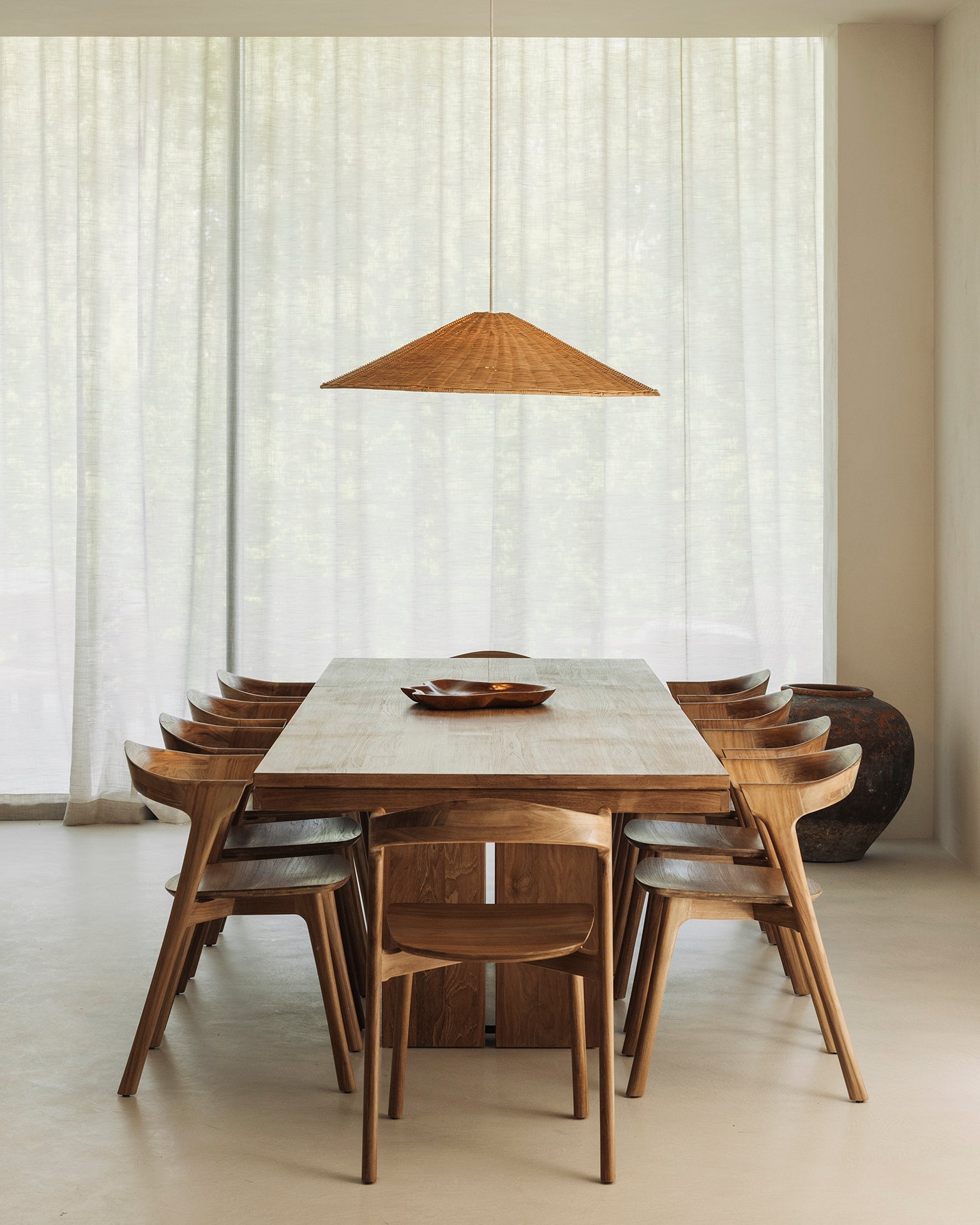 A minimalist dining room features the Ethnicraft Double Extendable Dining Table in natural teak, eight wooden chairs, a woven pendant light above, sheer white curtains, and a large ceramic pot in the background.
