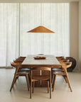 A minimalist dining room features the Ethnicraft Double Extendable Dining Table in natural teak, eight wooden chairs, a woven pendant light above, sheer white curtains, and a large ceramic pot in the background.