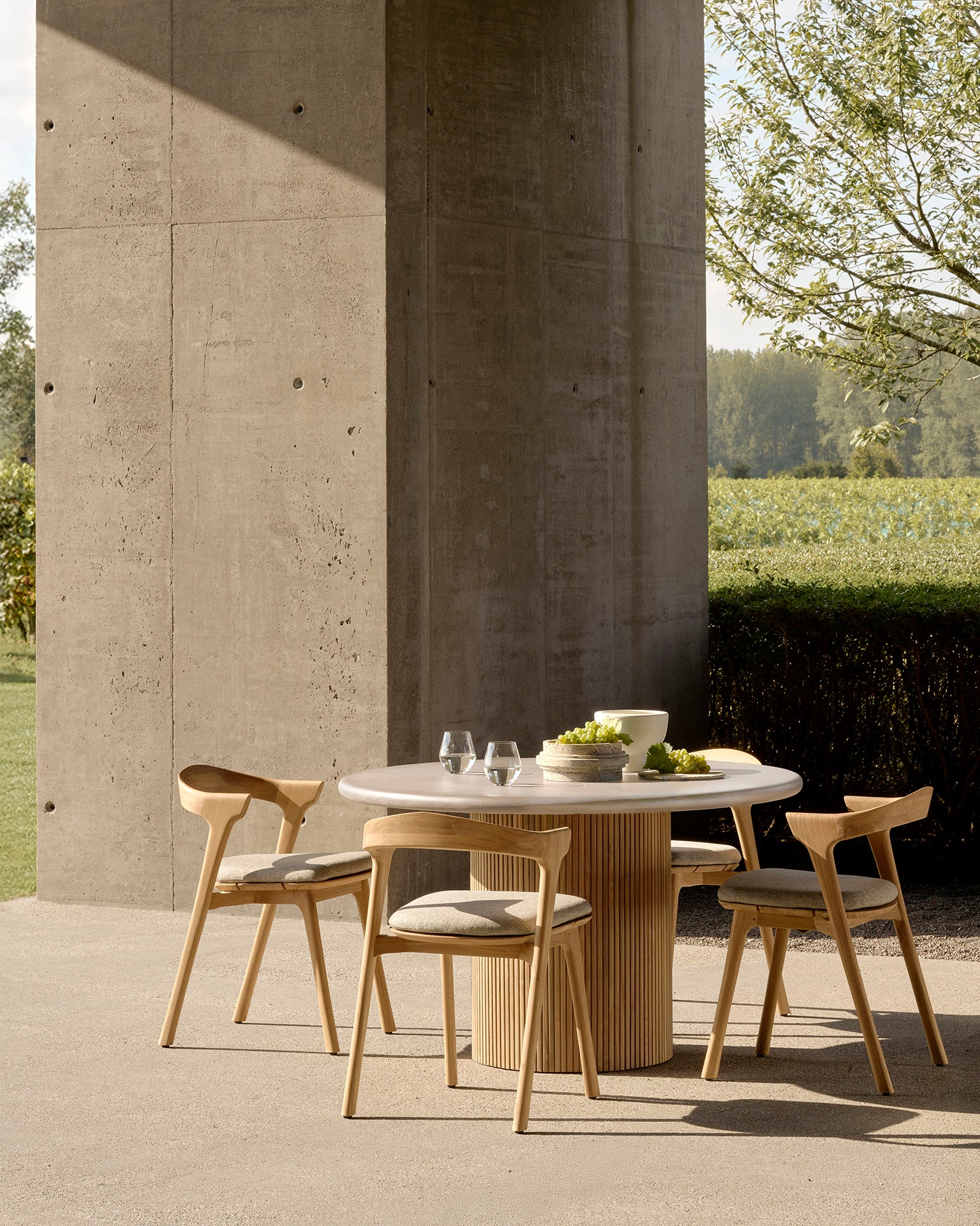 The Ethnicraft Roller Max Outdoor Dining Table with four matching chairs sits on a patio by a tall concrete wall, set with glassware and a bowl; crafted from reclaimed teak, it’s surrounded by lush greenery and trees.