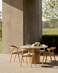 The Ethnicraft Roller Max Outdoor Dining Table with four matching chairs sits on a patio by a tall concrete wall, set with glassware and a bowl; crafted from reclaimed teak, it’s surrounded by lush greenery and trees.
