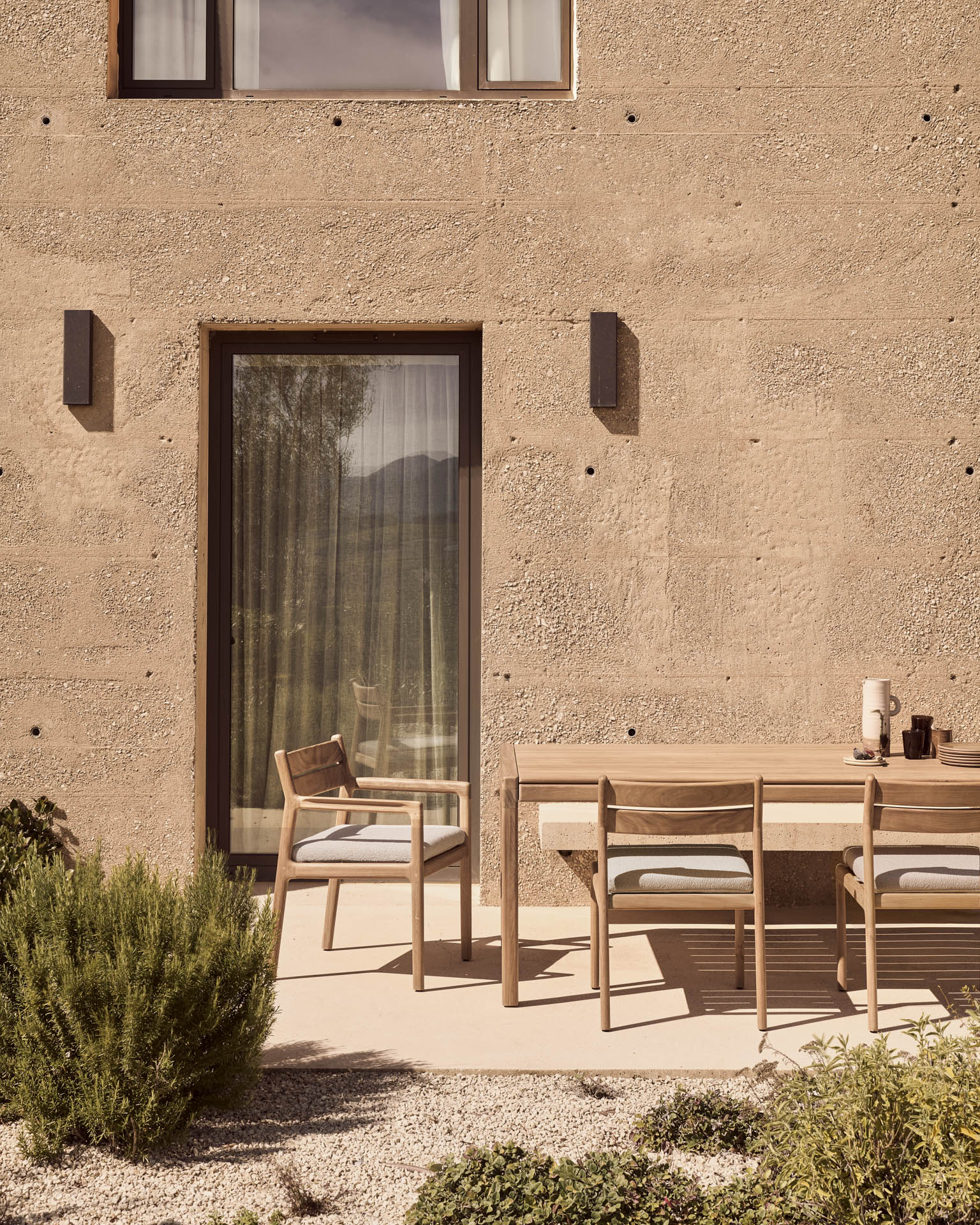 A minimalist outdoor patio showcases a solid teak dining table with three chairs, including a nearby Jack Outdoor Dining Chair - Teak (without armrest) by Ethnicraft, all surrounded by greenery in front of a textured beige wall with a glass door and window.