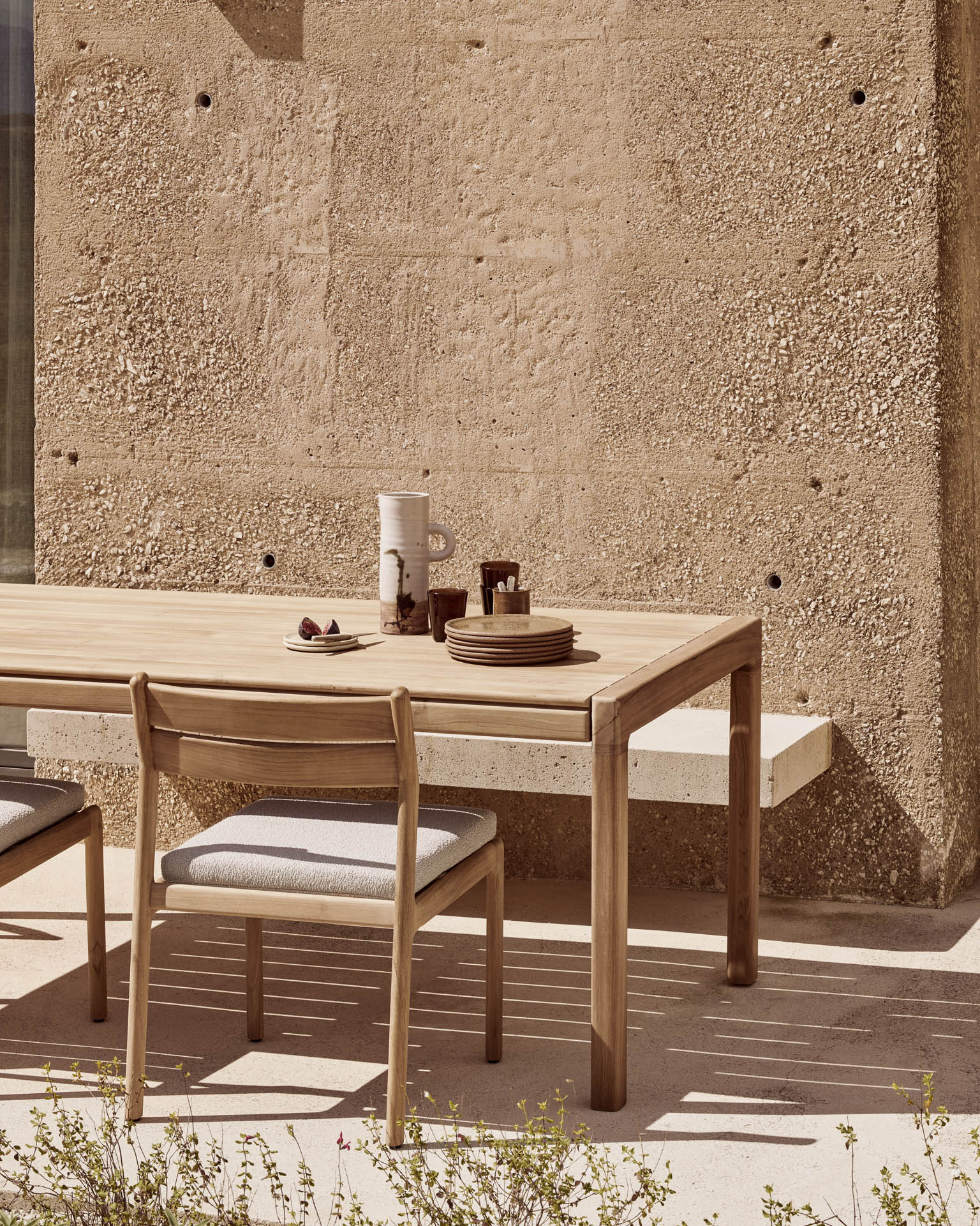 Two Jack Outdoor Dining Chairs - Teak (without armrest) by Ethnicraft are arranged around a wooden outdoor table set against a textured beige wall. Stacked plates, mugs, a white pitcher, and small bowls rest on the tabletop as sunlight casts soft shadows across the ground and wall.
