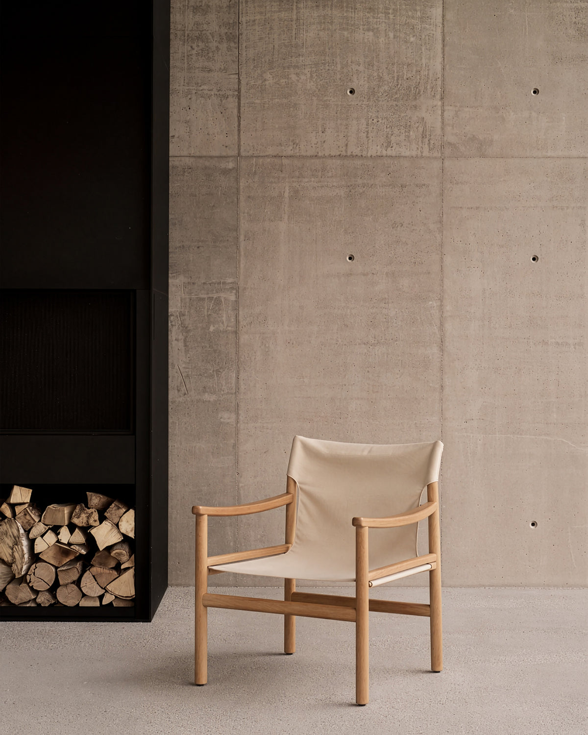 The Ethnicraft Safari Lounge Chair—a minimalist beige design echoing classic safari chairs with its wooden frame—rests on a light floor beside a neatly stacked pile of firewood, set against a concrete wall marked by visible grid lines and bolts.