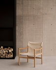 An Ethnicraft Safari Outdoor Lounge Chair with a beige seat cover sits next to a built-in black fireplace with stacked firewood, set against a textured concrete wall.