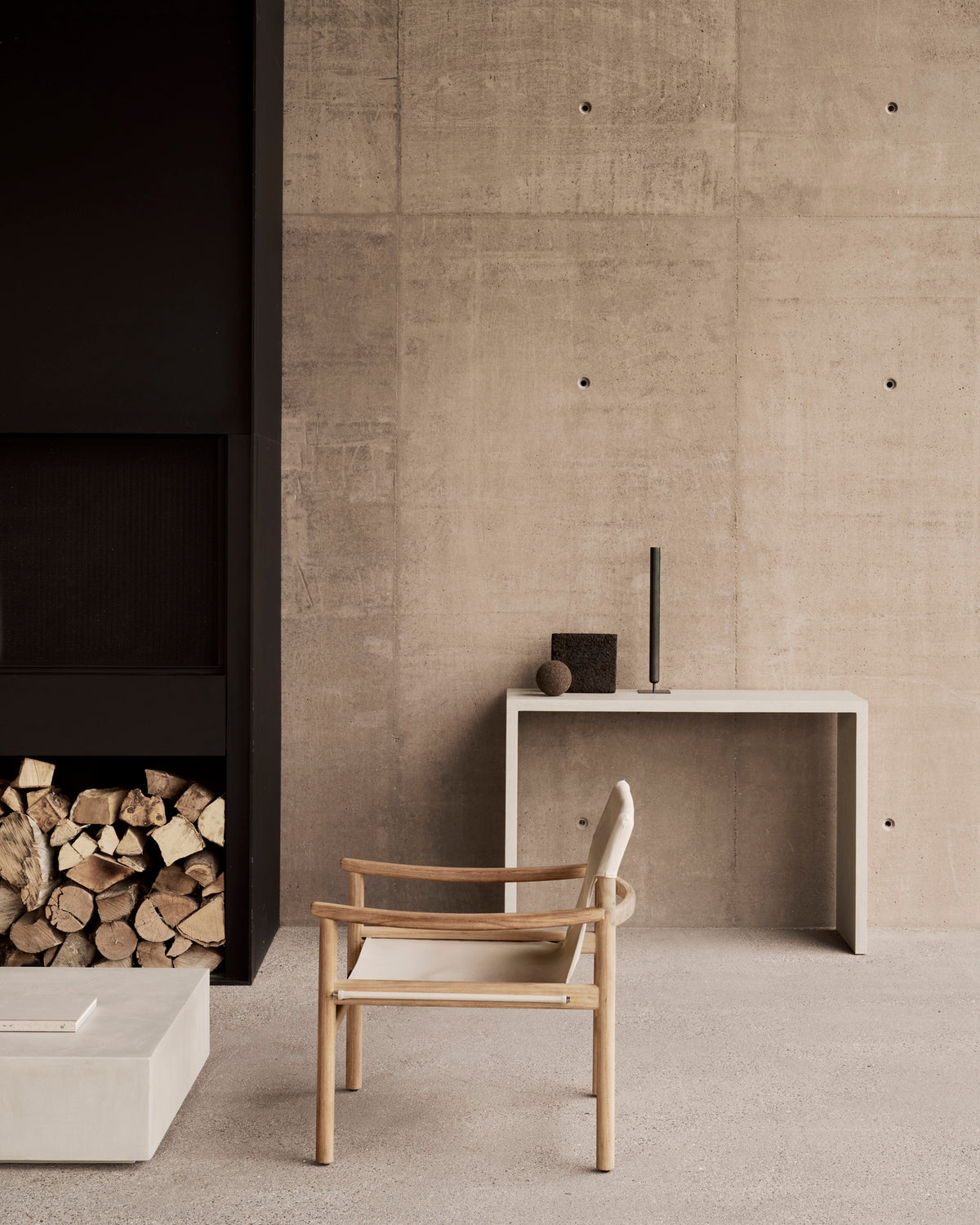 A minimalist room featuring the Ethnicraft Safari Lounge Chair in light wood, paired with a simple console table displaying decor items, a stack of firewood, and neutral-toned concrete walls and floor.