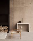 A minimalist room featuring the Ethnicraft Safari Lounge Chair in light wood, paired with a simple console table displaying decor items, a stack of firewood, and neutral-toned concrete walls and floor.