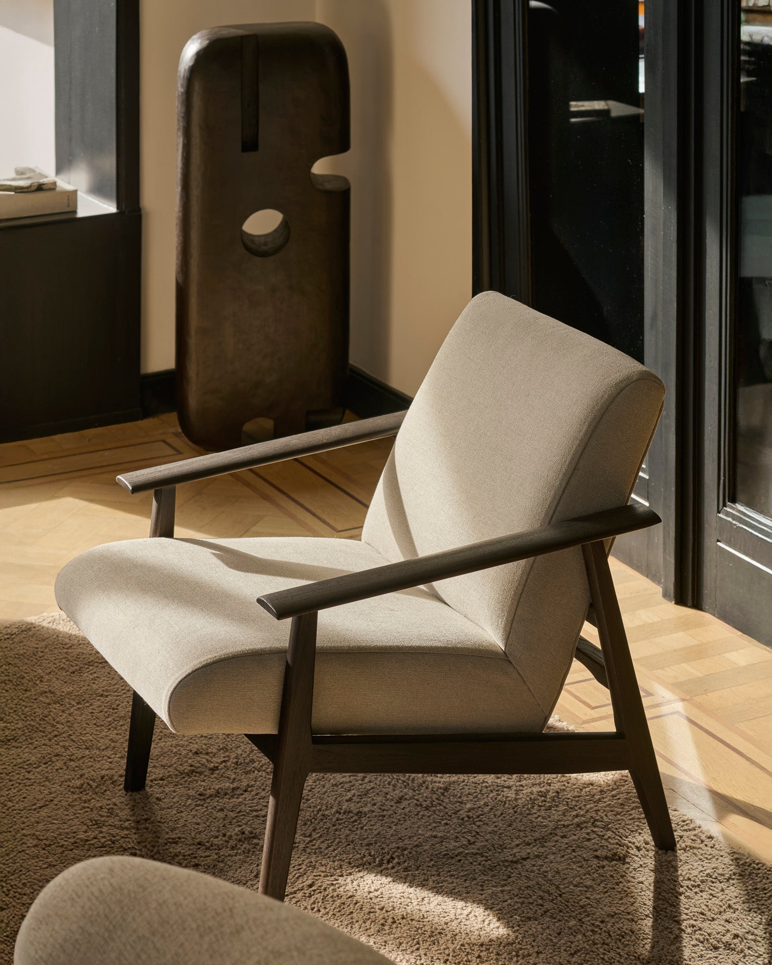 An Ethnicraft Aspekt Lounge Chair - Dark Brown Teak sits on a brown rug in a sunlit room with wooden floors. A sculptural object with round cutouts stands in the background near a black-framed glass door.