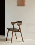 The Ethnicraft Bok Dining Chair - Teak Brown, with a curved backrest and angled legs, sits on light tiles against a minimalist beige wall. A rectangular piece of art or panel is mounted above the chair.
