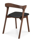 The Ethnicraft Bok Dining Chair - Teak Brown features a three-legged solid teak frame, curved backrest, and a black leather cushioned seat, displayed on a white background.
