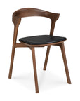 The Ethnicraft Bok Dining Chair - Teak Brown features a curved backrest, angled legs, and a black leather cushioned seat. Crafted from solid teak with a rich dark brown finish, it offers a sleek and contemporary look.