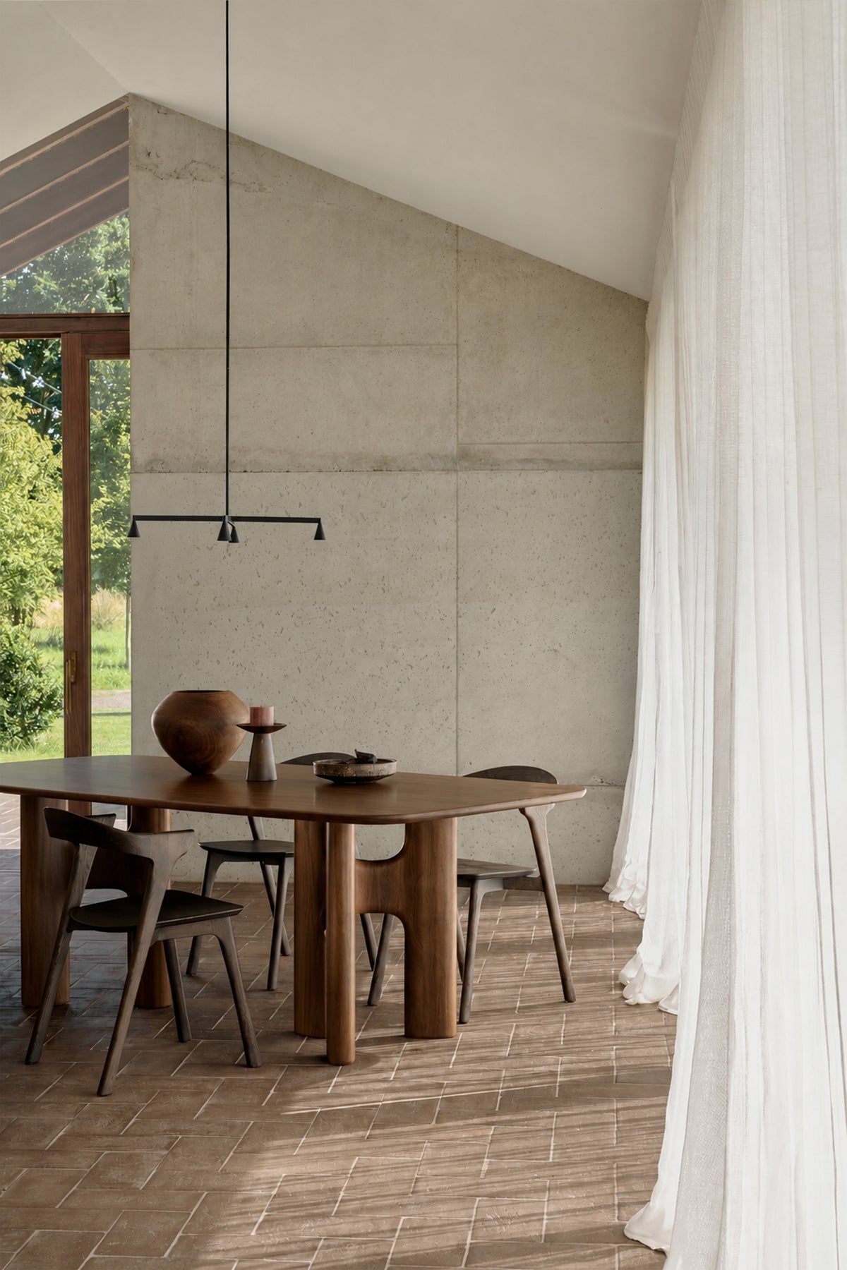 A dark solid wood Ethnicraft Flow Dining Table | Teak Wild Brown with rounded, cylindrical legs and four matching wooden chairs, set in a room with concrete walls and tiled flooring.