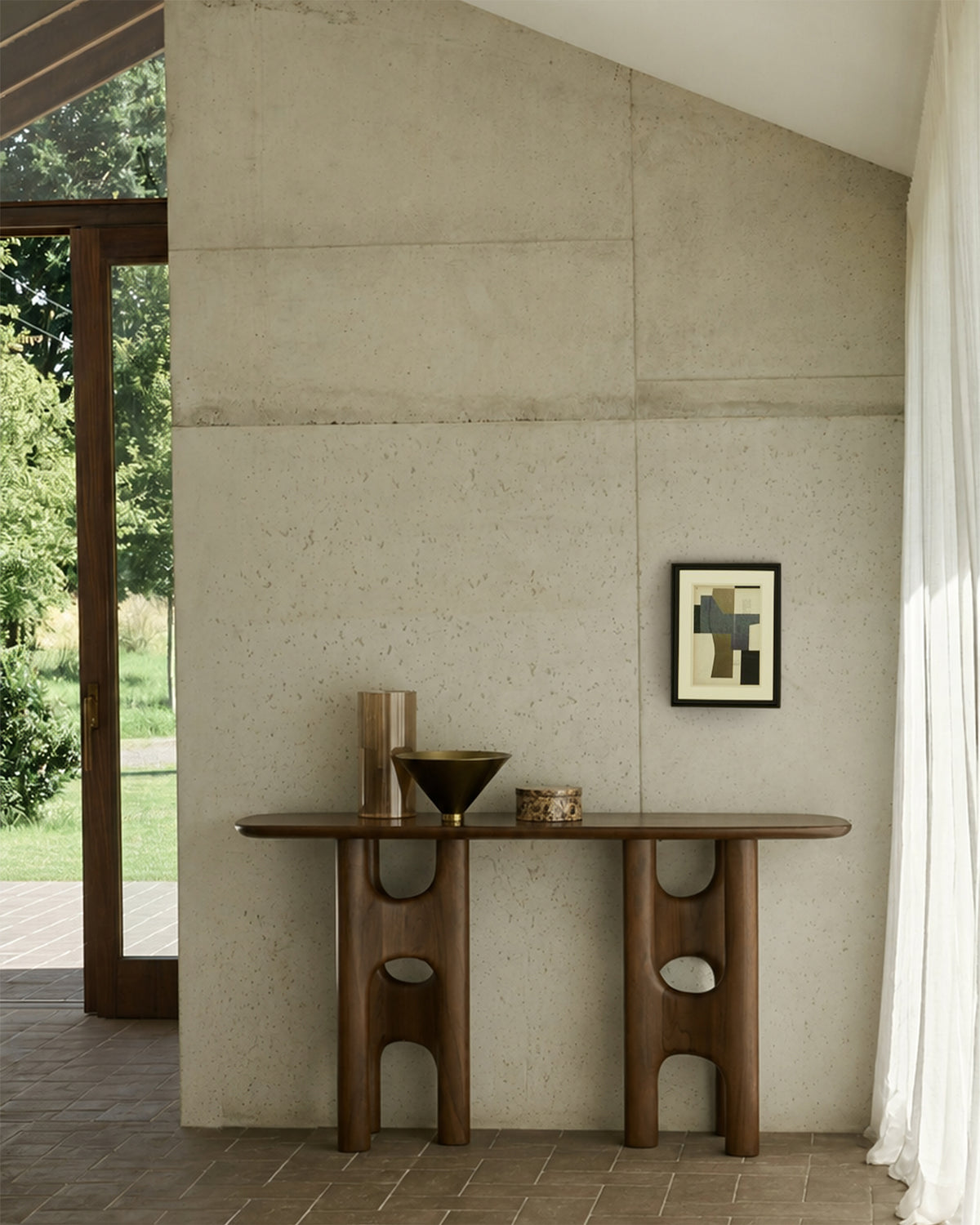 An Ethnicraft Flow Console | Teak Wild Brown with sculpted legs stands against a concrete wall, holding a vase, a bowl, and a small box beneath framed abstract art.