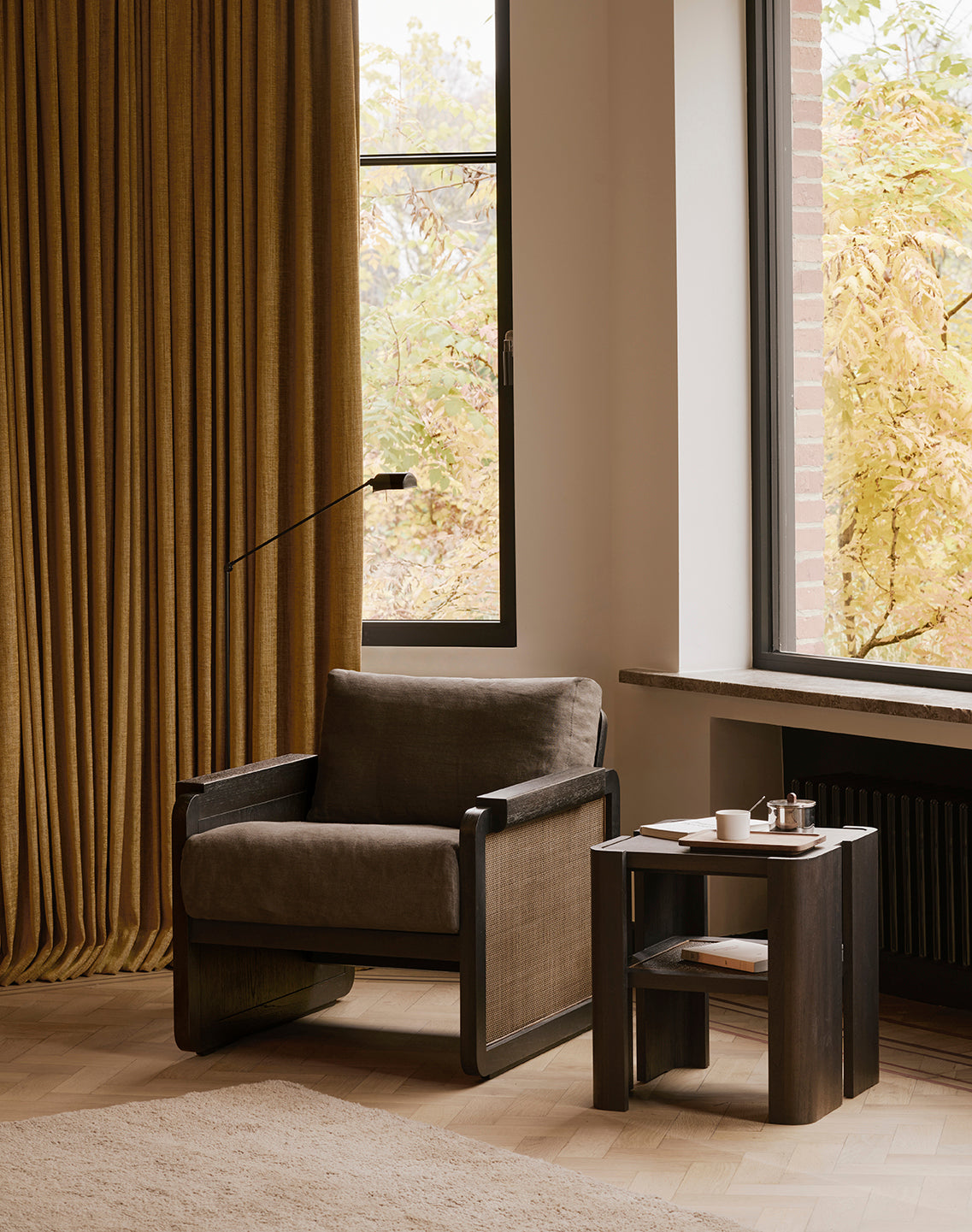 An Ethnicraft Panel Lounge Chair - Dark Brown Teak sits by a window with mustard curtains, next to a wooden side table holding a teapot, cup, and book. Autumn trees can be seen outside.